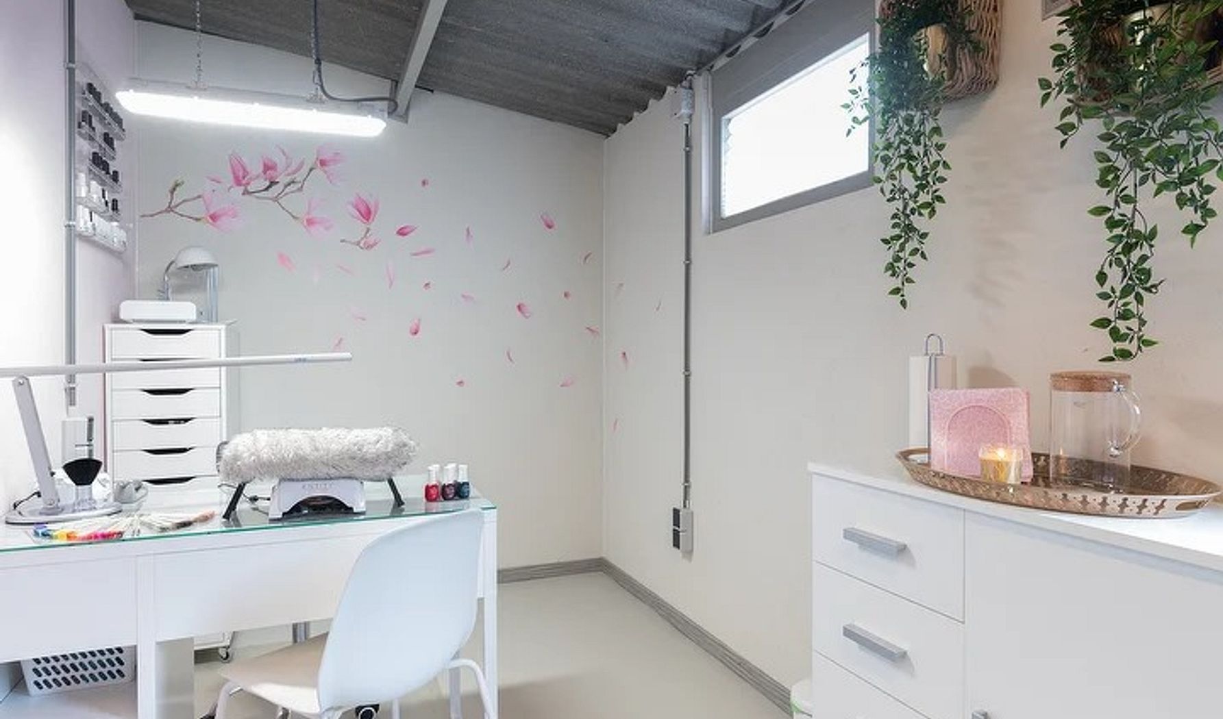 De salon: een witte ruimte met nageltafel, ledlamp, hangplanten en een geschilderde magnoliatak op de muur